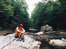 a woman sitting on a rock near a creek 