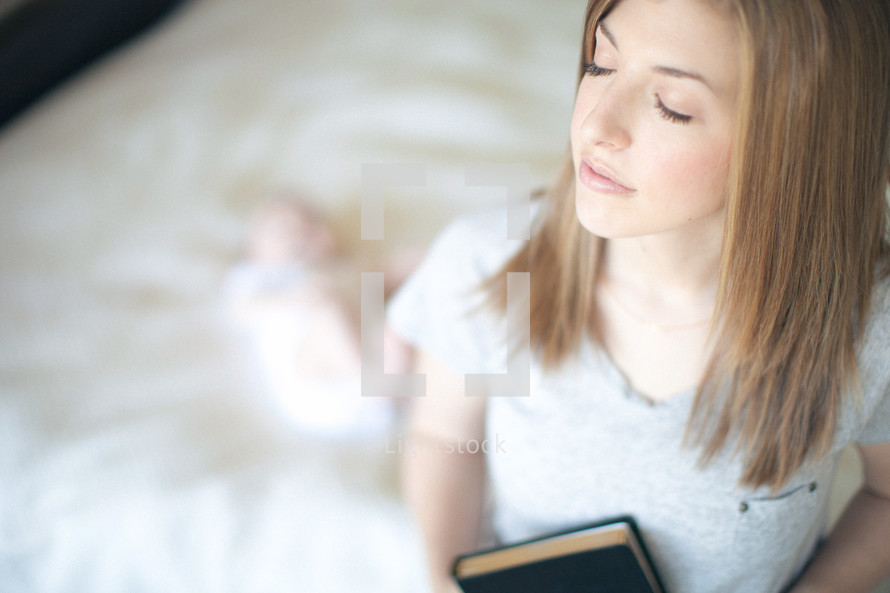 Woman with eyes closed holding Bible with infant daughter on the bed in the background.