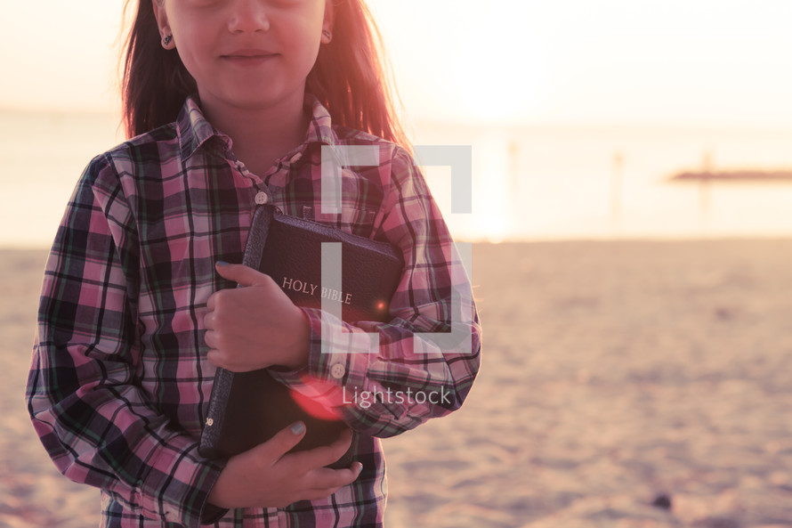 young girl happy, smiling, standing on the beach holding her bible at sunset