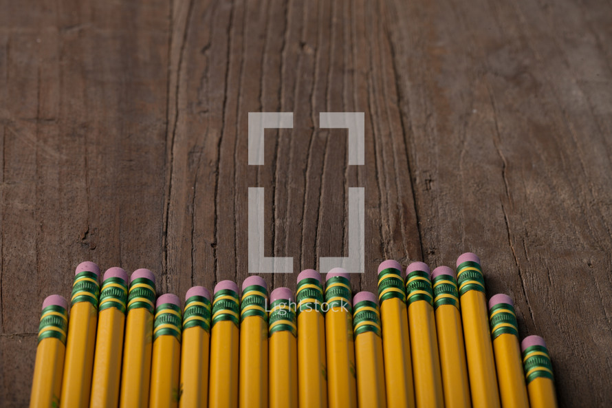 A row of yellow pencils with pink erasers on a wooden surface.