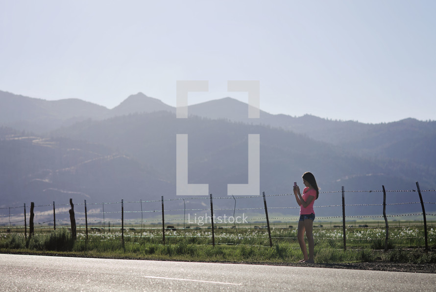a teen girl standing on the side of a rural road taking a picture with her cellphone 