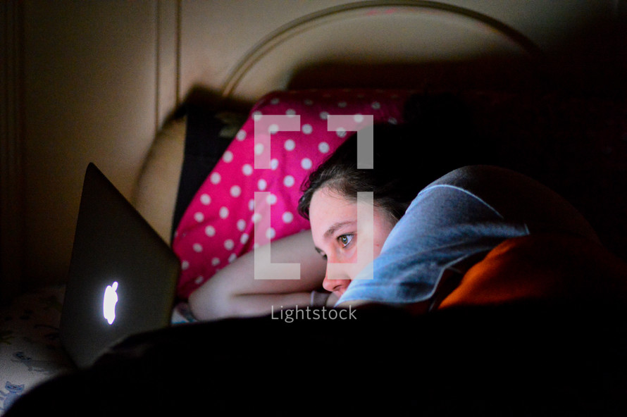 a girl lying in bed looking at her laptop 