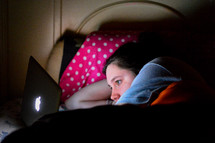 a girl lying in bed looking at her laptop 