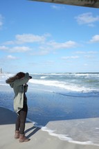 woman with a camera standing on a beach taking pictures 
