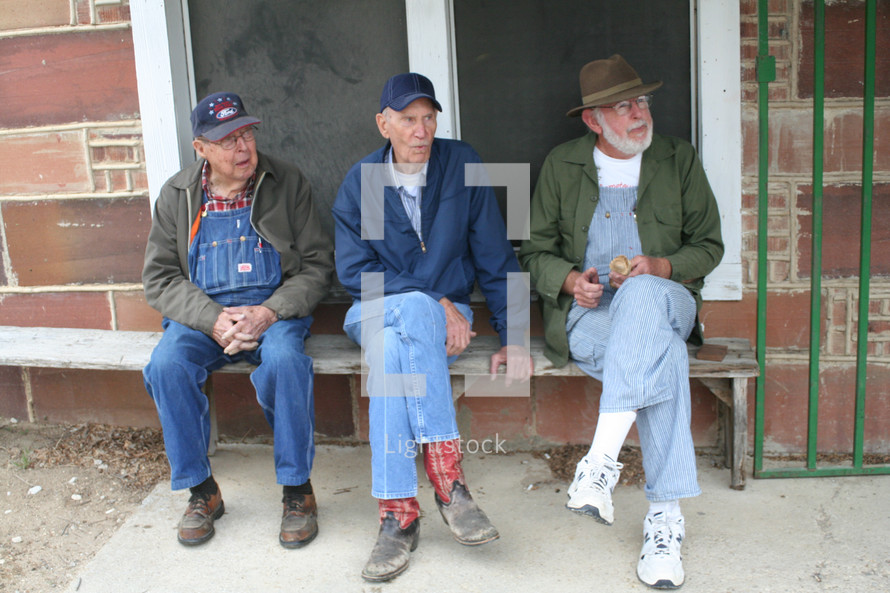 Three older men in overalls sitting outside on a bench.