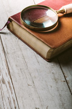 Magnifying glass on top of Bible laying on wooden table.