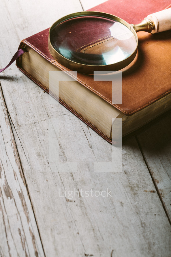 Magnifying glass on top of Bible laying on wooden table.