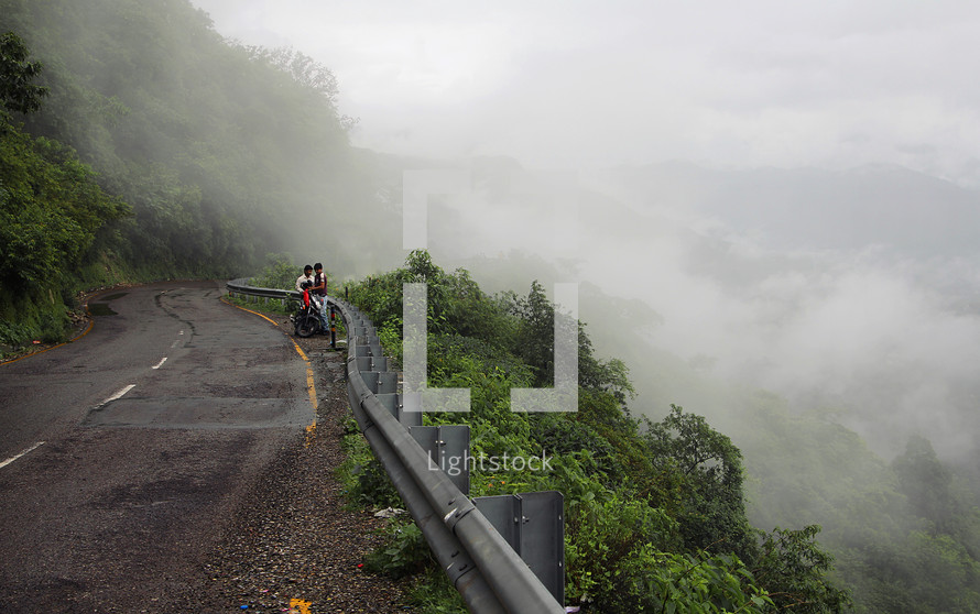 Two men with a motorcycle on the side of a winding mountain road.