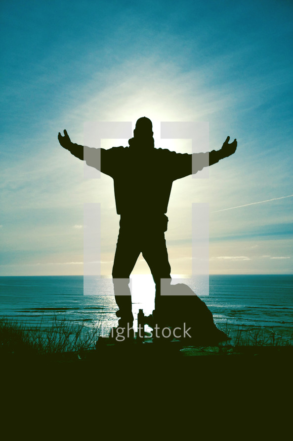 Silhouette of a man standing with outreached arms at the ocean with an aura of light around him.