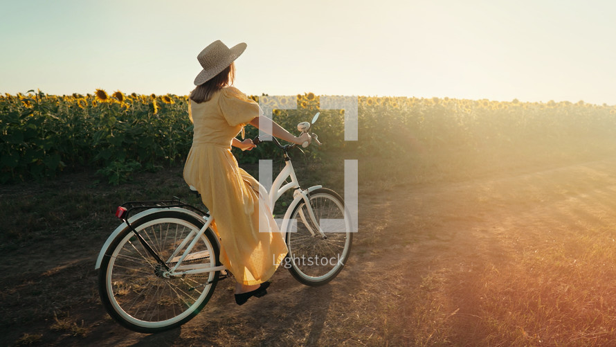 Rural woman in timeless dress riding retro styled white bicycle on country road alone near sunflowers field. Vintage fashion, amazing adventure, countryside activity, healthy lifestyle.