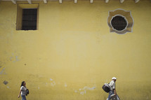 Two people walking next to a yellow wall.