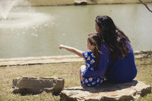 mother and daughter sitting on a rock looking out at a pond