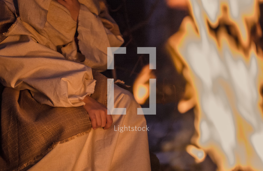 A woman in Biblical clothing sits near a fire.
