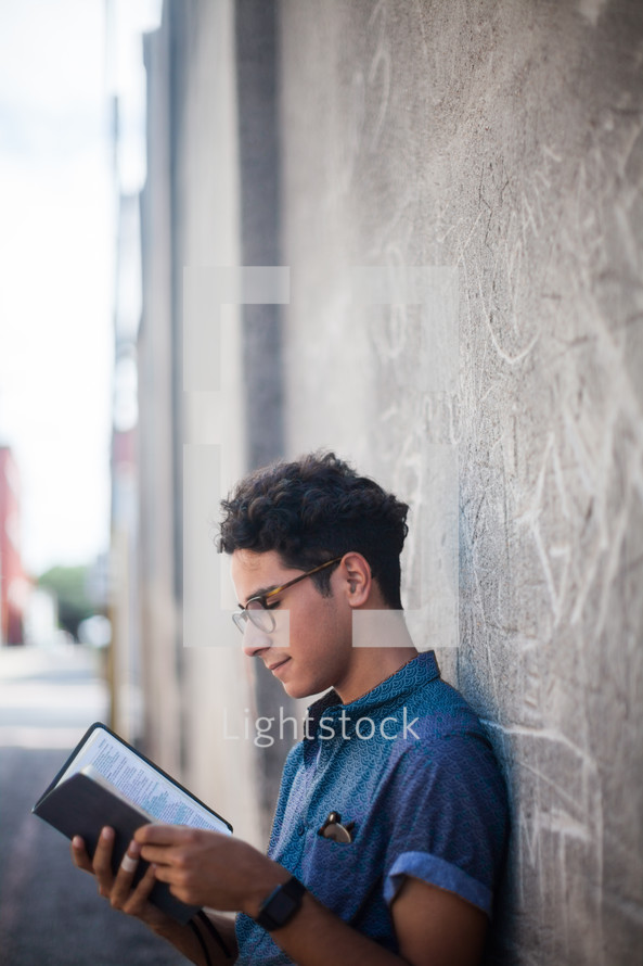 a man reading a Bible leaning against a gray wall 