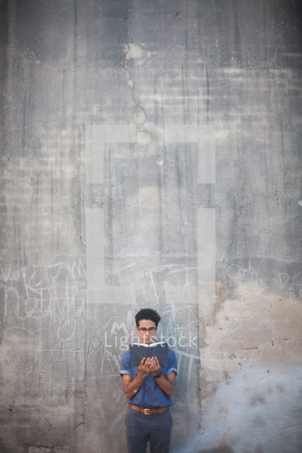 man reading a Bible in front of a gray wall with graffiti 