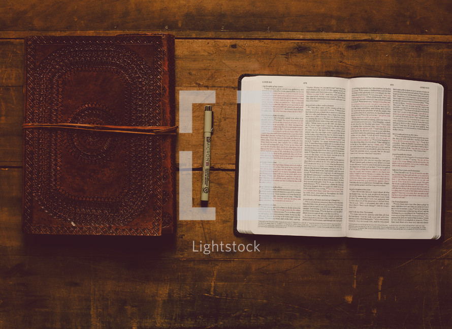 Open Bible, leather journal and a pen on a wooden table.