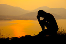 a silhouette of a woman kneeling in prayer by a lake 