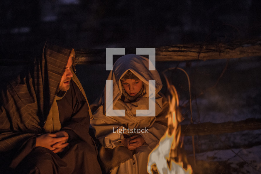 A man and child dressed in Biblical clothing sit near a fire.