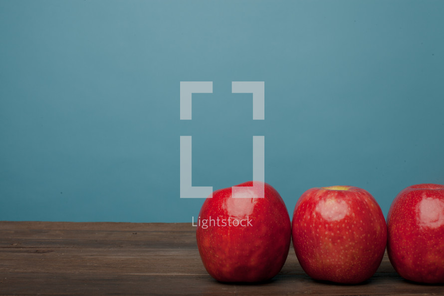 Three red apples lined up on a wooden surface with a blue background.