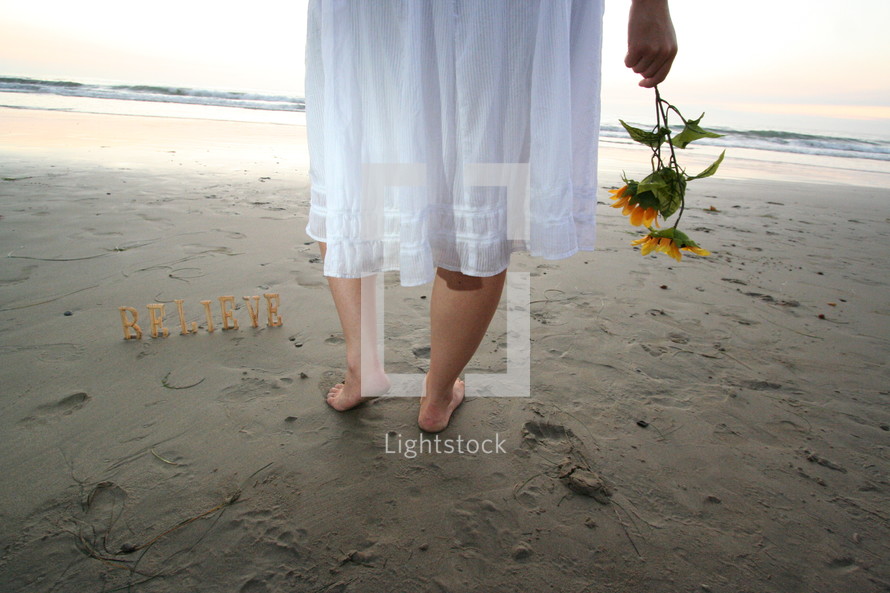 word BELIEVE on sand and woman in bare feet holding flowers