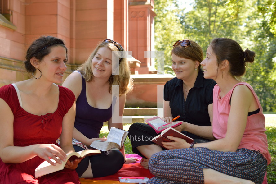 women's Bible study group having a discussion outdoors sitting on a blanket in the grass at a sunny summer day
