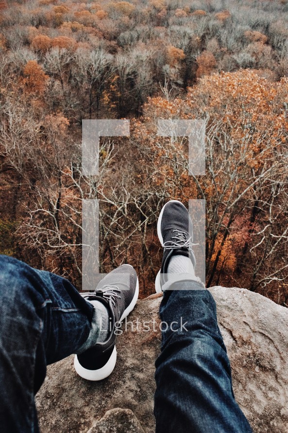 feet hanging off the side of a mountain over fall trees in a forest 
