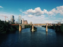 A railroad bridge over a river with city skyscrapers in the background.