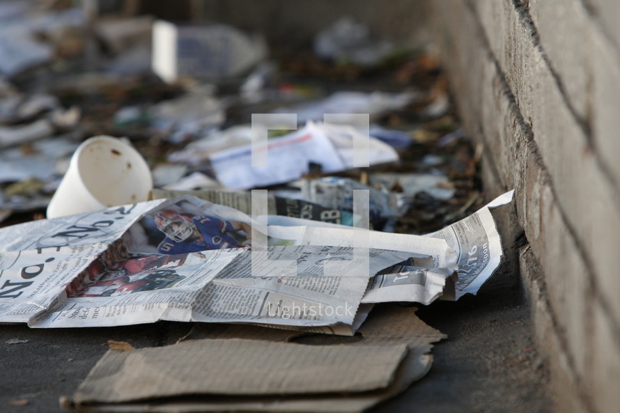 Newspaper and trash scattered on sidewalk against brick wall.