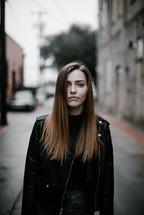 a woman in a leather jacket standing on a narrow city street 