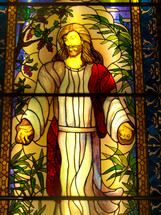 A gold, yellow, rose colored, red and brown Stained glass window of Jesus Christ, the son of God in a long white and red robe with his nail scarred hands stretched out and smiling peacefully showing grace, mercy and love to all who view this image.  