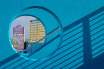 A brightly colored teal wall with a round window has a shadow from a metal railing