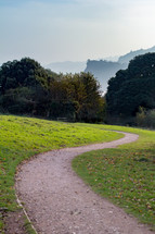 A trail winds through a green meadow towards a grove of trees with misty hills in the background