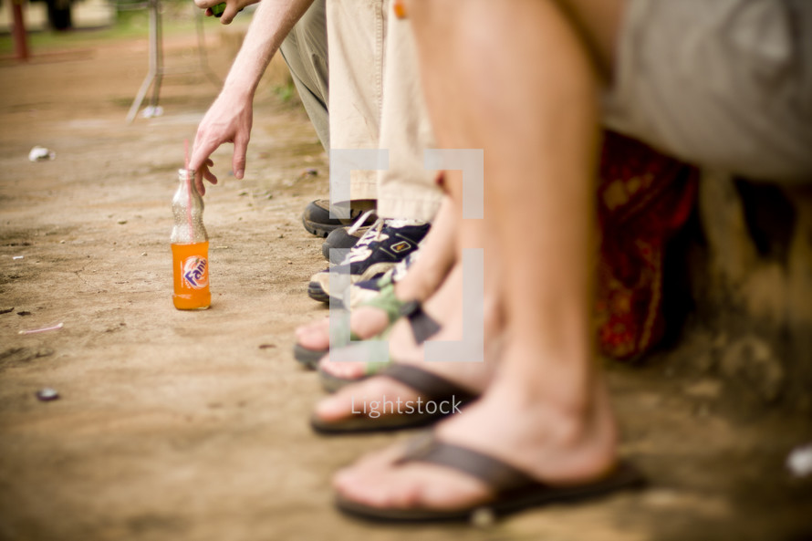 A row of legs and feet - A hand reaches for a drink