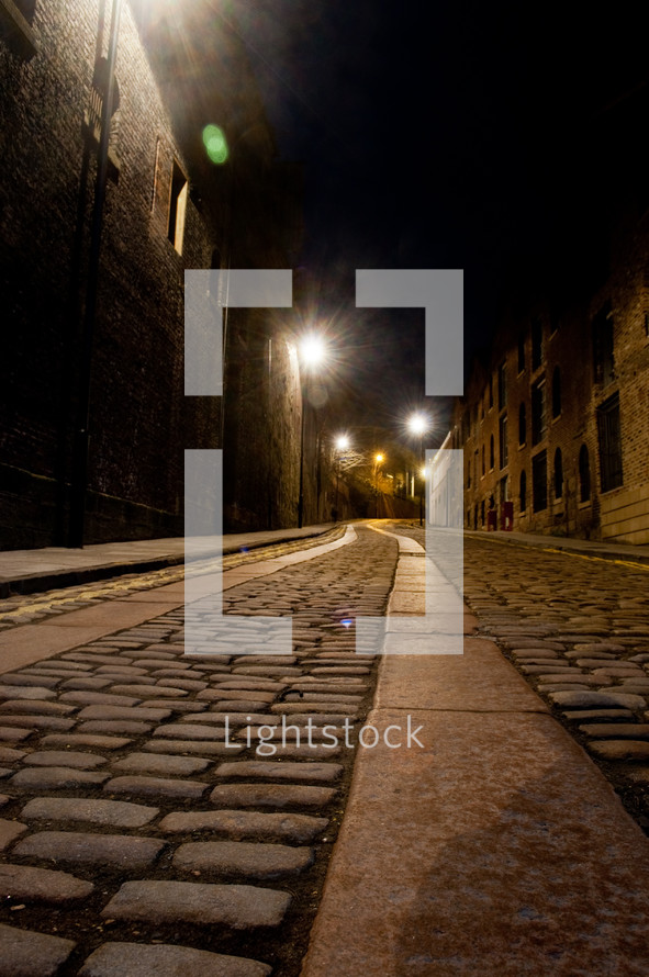 Cobblestone walkway in between brick buildings with beaming street lights in background.