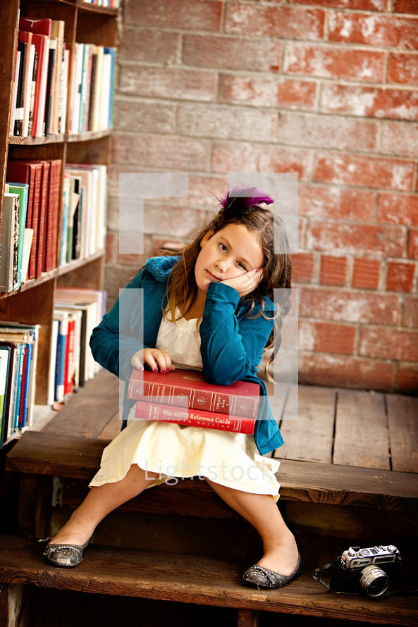 child sitting in a library with a stack of books in her lap and her head resting in her hand