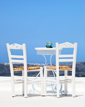 Two white chairs and a blurred panoramic view of the hills of santorini, Greece.