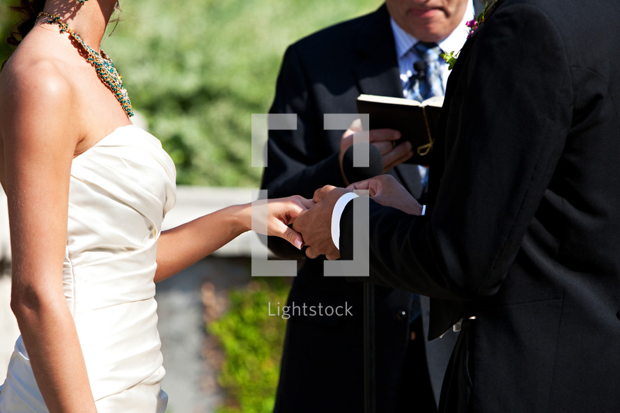 Groom placing ring on bride's hand during outdoor wedding ceremony.