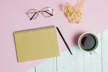 Writing concept with brown notebook, glasses, coffee cup and flowers over pink and wooden table. 