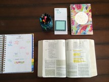 calendar, open Bible, journal, jar of pens, and notepad on a table 
