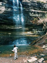 A boy stands watching a waterfall cascading down a cliff.