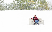 A man sits on a couch outside in the snow.