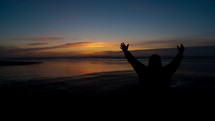 Person with arms raised by ocean at sunset.