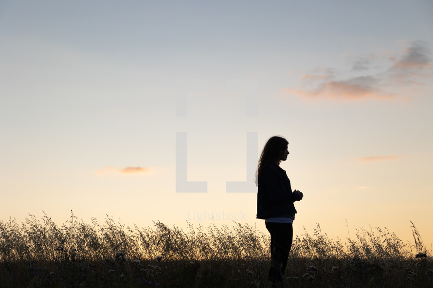 a teen girl praying outdoors in a field at sunset 