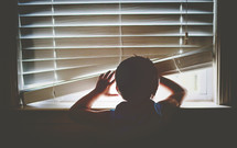 a child lifting window blinds and  looking through a window 