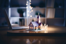 Miniature house on the table with a laptop and candles in the background