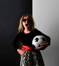 A young child in a fancy dress is holding a soccer sport ball and screaming for a girl's equality or girl power message