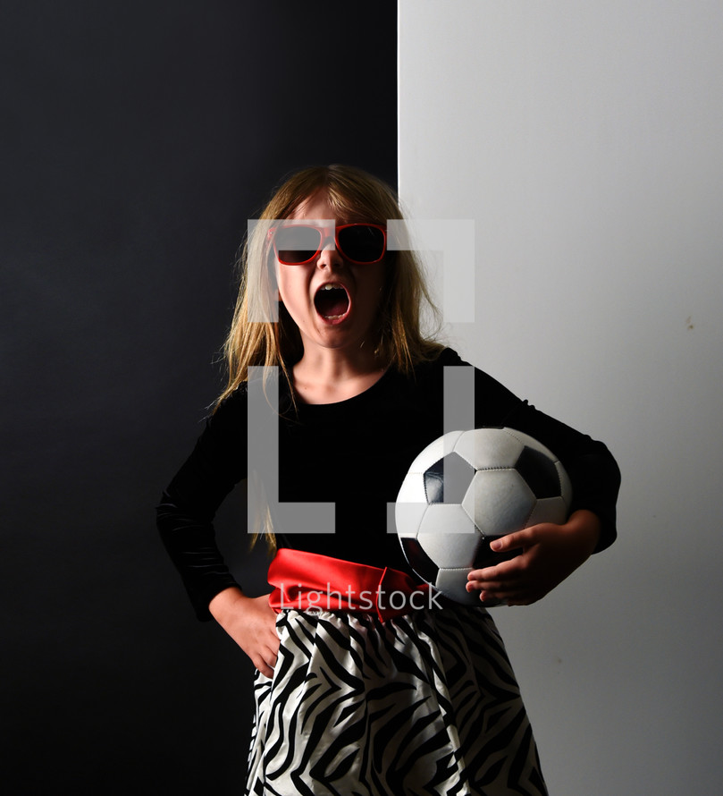A young child in a fancy dress is holding a soccer sport ball and screaming for a girl's equality or girl power message