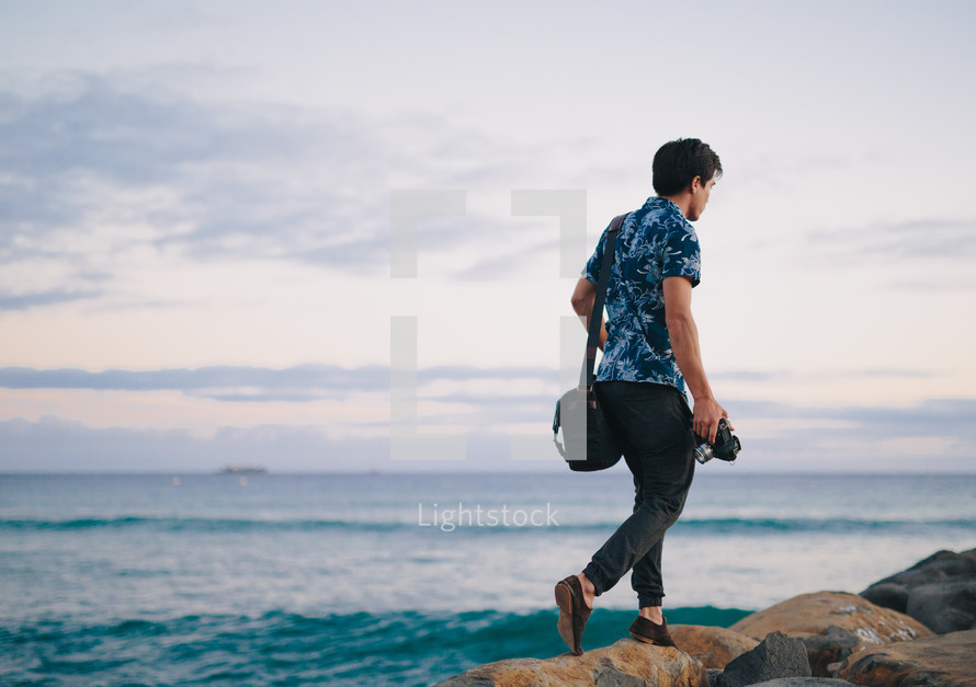man in a hawaiian shirt walking on rocks near the ocean carrying a camera 