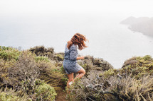 A woman walking through shrubberies toward the edge of a cliff above the ocean.