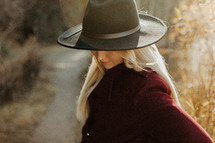a young woman in a hat and coat standing outdoors in golden sunlight 